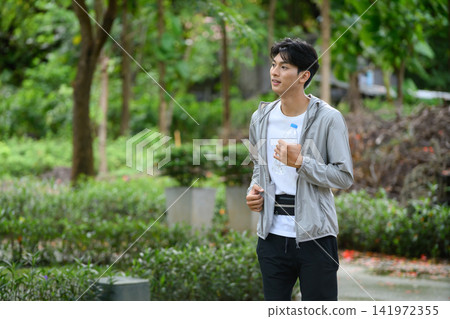 Young Asian Man Jogging in Park Holding Water Bottle with Green Nature Background 141972355
