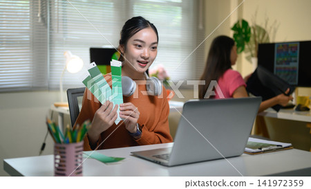 Young Asian Female Creative Designer Holding Green Color Swatches at Office Desk with Laptop Young Asian Female Creative Designer Holding Green Color Swatches at Office Desk with Laptop 141972359