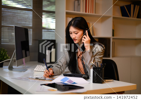Young Asian Businesswoman Talking on Phone While Taking Notes at Office Desk 141972452