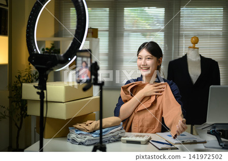 Young Asian Woman Live Streaming Fashion Sales from Home Office with Ring Light and Smartphone Young Asian Woman Live Streaming Fashion Sales from Home Office with Ring Light and Smartphone 141972502