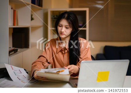 Young Asian Businesswoman Reviewing Documents and Reports at Office Desk with Laptop Young Asian Businesswoman Reviewing Documents and Reports at Office Desk with Laptop 141972553