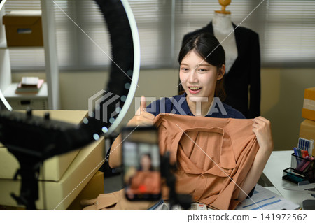 Young Asian Woman Live Streaming Fashion Clothing Sale with Ring Light and Smartphone for Small Business Young Asian Woman Live Streaming Fashion Clothing Sale with Ring Light and Smartphone for Small Business 141972562