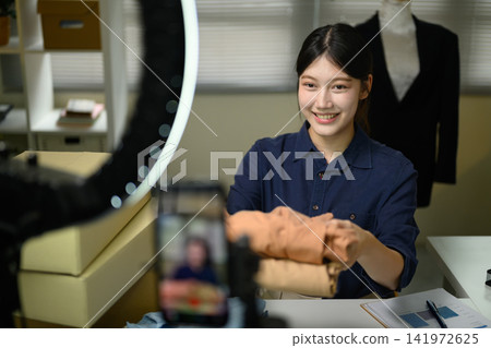 Young Asian Woman Live Streaming Fashion Clothing Sales with Ring Light and Smartphone Camera for Small Business Young Asian Woman Live Streaming Fashion Clothing Sales with Ring Light and Smartphone Camera for Small Business 141972625
