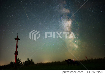 Milky Way and Meteor Shower over Wooden Wayside Cross and People Relaxing under Starry Night Sky Milky Way and Meteor Shower over Wooden Wayside Cross and People Relaxing under Starry Night Sky 141972950