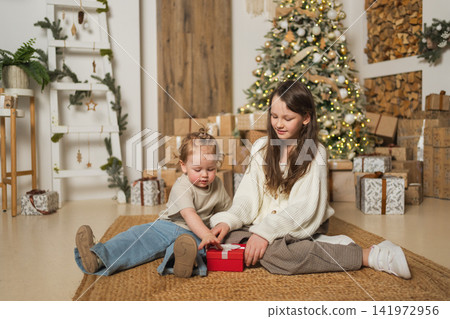 Merry Christmas Xmas holiday. Children sisters girls unpacking open up gift box near Christmas tree. Kids in living room with Christmas tree opening gift box with surprise faces. Christmas eve at home Merry Christmas Xmas holiday. Children sisters girls unpacking open up gift box near Christmas tree. Kids in living room with Christmas tree opening gift box with surprise faces. Christmas eve at home 141972956