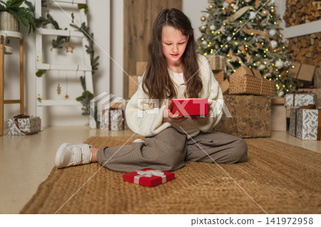 Merry Christmas Xmas holiday. Teenage girl unpacking open up gift box near Christmas tree. Teen kid child in living room with Christmas tree opening gift box with surprise face. Christmas eve at home Merry Christmas Xmas holiday. Teenage girl unpacking open up gift box near Christmas tree. Teen kid child in living room with Christmas tree opening gift box with surprise face. Christmas eve at home 141972958