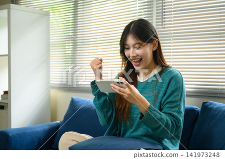 Excited Young Asian Woman Celebrating Good News on Smartphone While Sitting on Sofa at Home Excited Young Asian Woman Celebrating Good News on Smartphone While Sitting on Sofa at Home 141973248