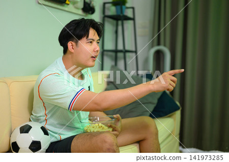 Excited Asian Man Cheering Football Game at Home Sitting on Sofa with Soccer Ball and Popcorn Excited Asian Man Cheering Football Game at Home Sitting on Sofa with Soccer Ball and Popcorn 141973285