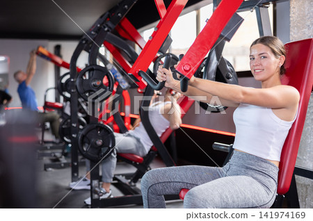 Girl doing strength training in gym, exercising upper body muscles on shoulder press machine 141974169