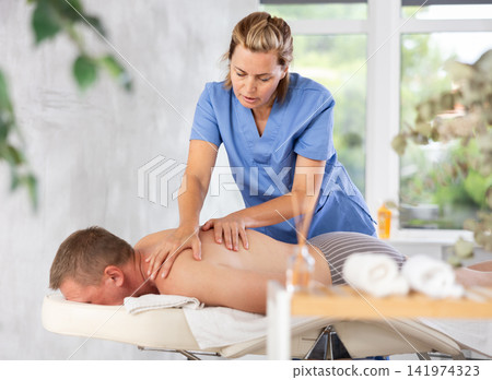 Female masseur performs back stroking and patting during massage session for male client Female masseur performs back stroking and patting during massage session for male client 141974323