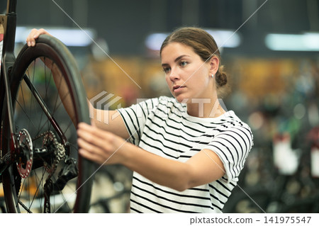 Positive young girl looking for and trying wheel new bike in sports bicycle store Positive young girl looking for and trying wheel new bike in sports bicycle store 141975547