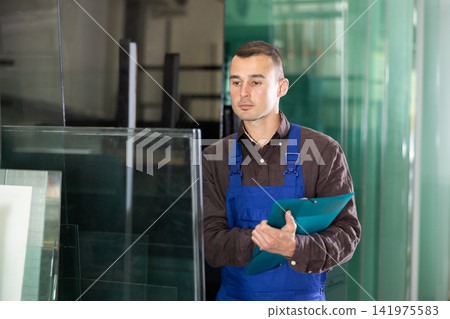 Experienced worker checks the quality of made window glass in an industrial workshop, marks the data in documents 141975583