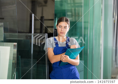Female worker checks the quality of made window glass in an industrial workshop, marks the data in documents Female worker checks the quality of made window glass in an industrial workshop, marks the data in documents 141975597