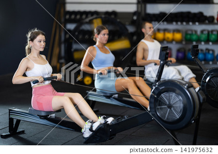 Athletic woman in sportswear training on rowing machine in sport gym Athletic woman in sportswear training on rowing machine in sport gym 141975632
