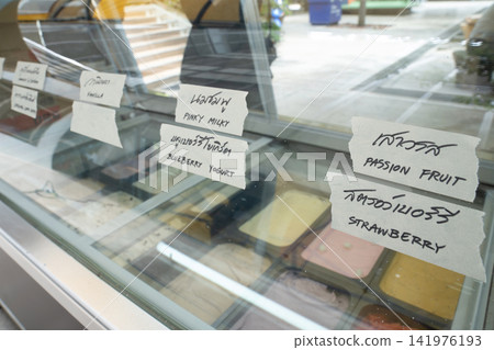 Glass ice cream freezer with handwritten Thai labels and English translations for assorted flavors including taro, coffee, vanilla, yogurt, passion fruit, and strawberry. 141976193