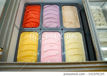 Various colorful ice cream flavors are neatly arranged in metal tubs inside a commercial freezer display. Rows of freshly churned gelato offer sweet dessert choices. Various colorful ice cream flavors are neatly arranged in metal tubs inside a commercial freezer display. Rows of freshly churned gelato offer sweet dessert choices. 141976195