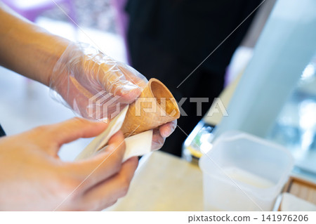 A worker wearing plastic gloves wraps a napkin around a waffle cone. 141976206