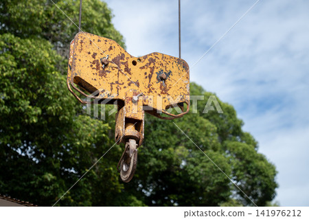 An old yellow crane sheave block with a steel hook hangs outdoors. An old yellow crane sheave block with a steel hook hangs outdoors. 141976212