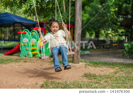 happy preschool child girl swinging on a wooden swing in a playground. happy preschool child girl swinging on a wooden swing in a playground. 141976300