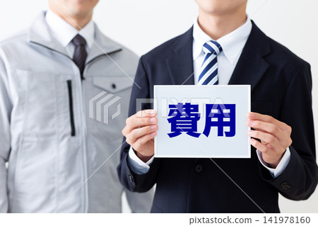 一位身著西裝的年輕商人,和一位身穿工作服、手持寫有成本的白板的中年男子。 一位身著西裝的年輕商人,和一位身穿工作服、手持寫有成本的白板的中年男子。 141978160