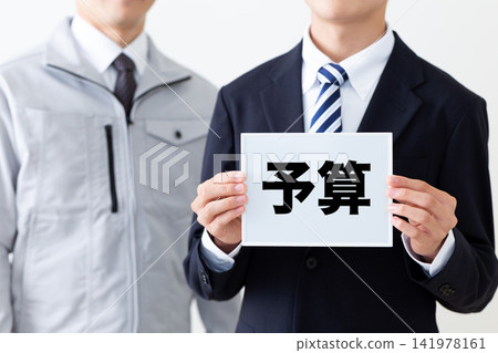 一位身穿西裝、手持預算白板的年輕商人,以及一位身穿工作服的中年男子。 一位身穿西裝、手持預算白板的年輕商人,以及一位身穿工作服的中年男子。 141978161