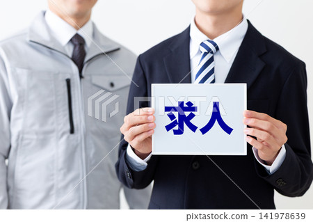 A young businessman in a suit holding a whiteboard advertising job openings, and a middle-aged man in work clothes. A young businessman in a suit holding a whiteboard advertising job openings, and a middle-aged man in work clothes. 141978639