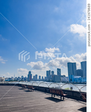 A wooden deck bench by the riverside, with a blue sky and clouds stretching out above, and a cluster of skyscrapers in the background. A wooden deck bench by the riverside, with a blue sky and clouds stretching out above, and a cluster of skyscrapers in the background. 141978689