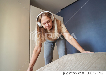 Woman Leaning Forward Arranging Blanket Edges While Wearing Headphones In Compact Bedroom With Blue Woman Leaning Forward Arranging Blanket Edges While Wearing Headphones In Compact Bedroom With Blue 141979742