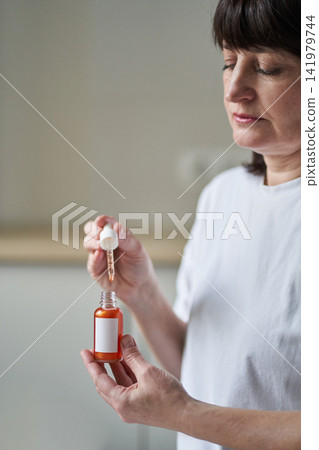 Middle-Aged Woman Demonstrating Serum Dropper Over Bottle While Looking Down In Home Kitchen Middle-Aged Woman Demonstrating Serum Dropper Over Bottle While Looking Down In Home Kitchen 141979744