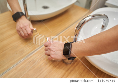 Person Turning Bathroom Faucet Knob At Sink Wearing Smartwatch Reflected In Mirror On Wooden Person Turning Bathroom Faucet Knob At Sink Wearing Smartwatch Reflected In Mirror On Wooden 141979745