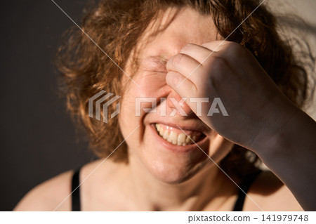 Woman Covering Eyes With Hand And Smiling In Warm Indoor Close Up Portrait Under Directional Light Woman Covering Eyes With Hand And Smiling In Warm Indoor Close Up Portrait Under Directional Light 141979748