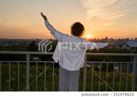 Person With Arms Raised At Railing During Sunset Above City Skyline And Grassy Overlook In Open Person With Arms Raised At Railing During Sunset Above City Skyline And Grassy Overlook In Open 141979828