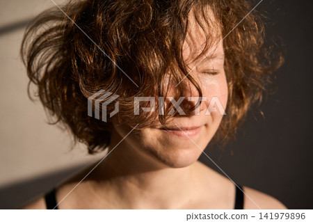Young Woman With Curly Hair Smiling Softly In Warm Indoor Sunlight Near Plain Wall Young Woman With Curly Hair Smiling Softly In Warm Indoor Sunlight Near Plain Wall 141979896