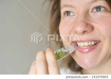 Young Woman Smiling While Applying Lip Gloss From Glass Roller Bottle In Bright Neutral Indoor Young Woman Smiling While Applying Lip Gloss From Glass Roller Bottle In Bright Neutral Indoor 141979905