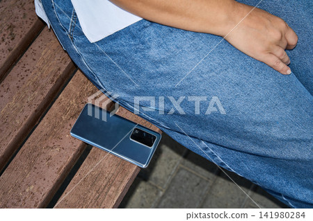 Smartphone Resting On Wooden Bench Beside Seated Woman In Denim Outfit At Park Walkway With Close Smartphone Resting On Wooden Bench Beside Seated Woman In Denim Outfit At Park Walkway With Close 141980284