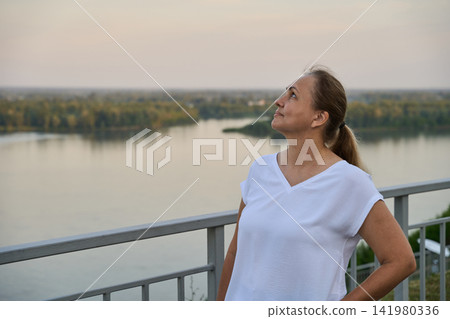 Adult Woman Standing Beside Railing And Looking Upward Over River Landscape In Soft Evening Light 141980336