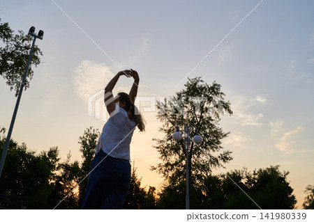 Woman Stretching Arms Overhead In Side Silhouette At Sunset Among Trees And Park Lights Woman Stretching Arms Overhead In Side Silhouette At Sunset Among Trees And Park Lights 141980339