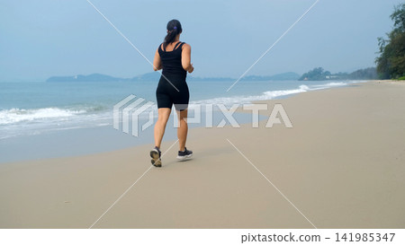 Rear view of young woman in black athletic outfit running steadily along a flat sandy beach with open ocean and misty hills behind. Fitness outdoor concept Rear view of young woman in black athletic outfit running steadily along a flat sandy beach with open ocean and misty hills behind. Fitness outdoor concept 141985347