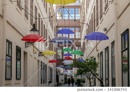 Colorful hanging umbrellas decorate a narrow historic alley with distant pedestrians Colorful hanging umbrellas decorate a narrow historic alley with distant pedestrians 141986743