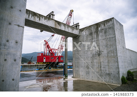 Under a cloudy sky, a large red and white crane ship and the harbor scenery can be seen through an opening in the brand new seawall. Under a cloudy sky, a large red and white crane ship and the harbor scenery can be seen through an opening in the brand new seawall. 141988373