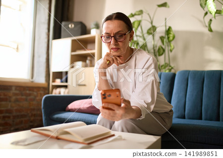 Focused mature woman checking mobile payment details at home Focused mature woman checking mobile payment details at home 141989851