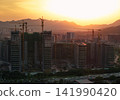 Tall residential buildings under construction with multiple cranes rising above the city skyline during a golden sunset near mountains in China 141990420