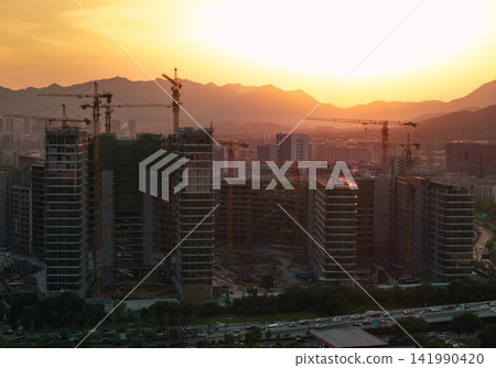 Tall residential buildings under construction with multiple cranes rising above the city skyline during a golden sunset near mountains in China Tall residential buildings under construction with multiple cranes rising above the city skyline during a golden sunset near mountains in China 141990420