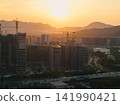 Tall residential buildings under construction with multiple cranes rising above the city skyline during a golden sunset near mountains in China 141990421