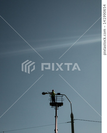 Electrician fixing street lamp in bucket truck platform against blue sky Electrician fixing street lamp in bucket truck platform against blue sky 141990654