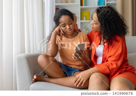 Concerned Black mother comforting her upset daughter while she looking at smartphone Concerned Black mother comforting her upset daughter while she looking at smartphone 141990678