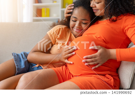 Closeup of black daughter resting against pregnant mothers belly, both smiling Closeup of black daughter resting against pregnant mothers belly, both smiling 141990692