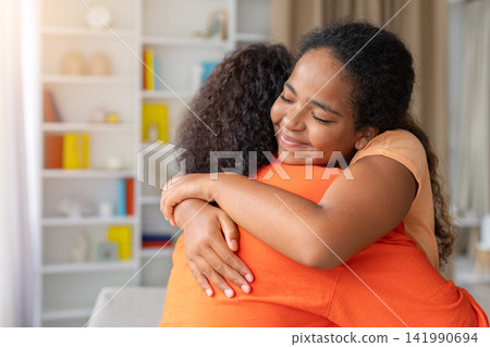 Young Black daughter hugging her mother tightly at home with peaceful smile Young Black daughter hugging her mother tightly at home with peaceful smile 141990694