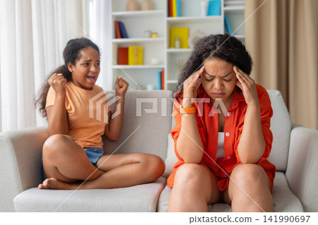 Stressed black mother holding head in pain while daughter shouts angrily behind Stressed black mother holding head in pain while daughter shouts angrily behind 141990697