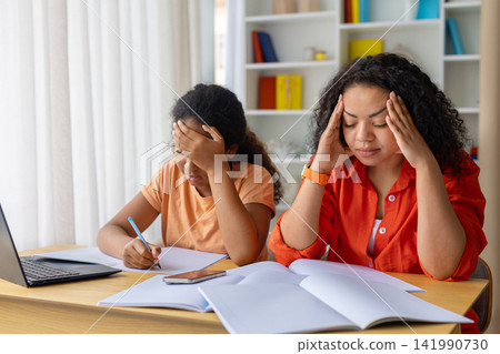 Stressed Black mother and daughter sitting at desk with books and notebooks Stressed Black mother and daughter sitting at desk with books and notebooks 141990730